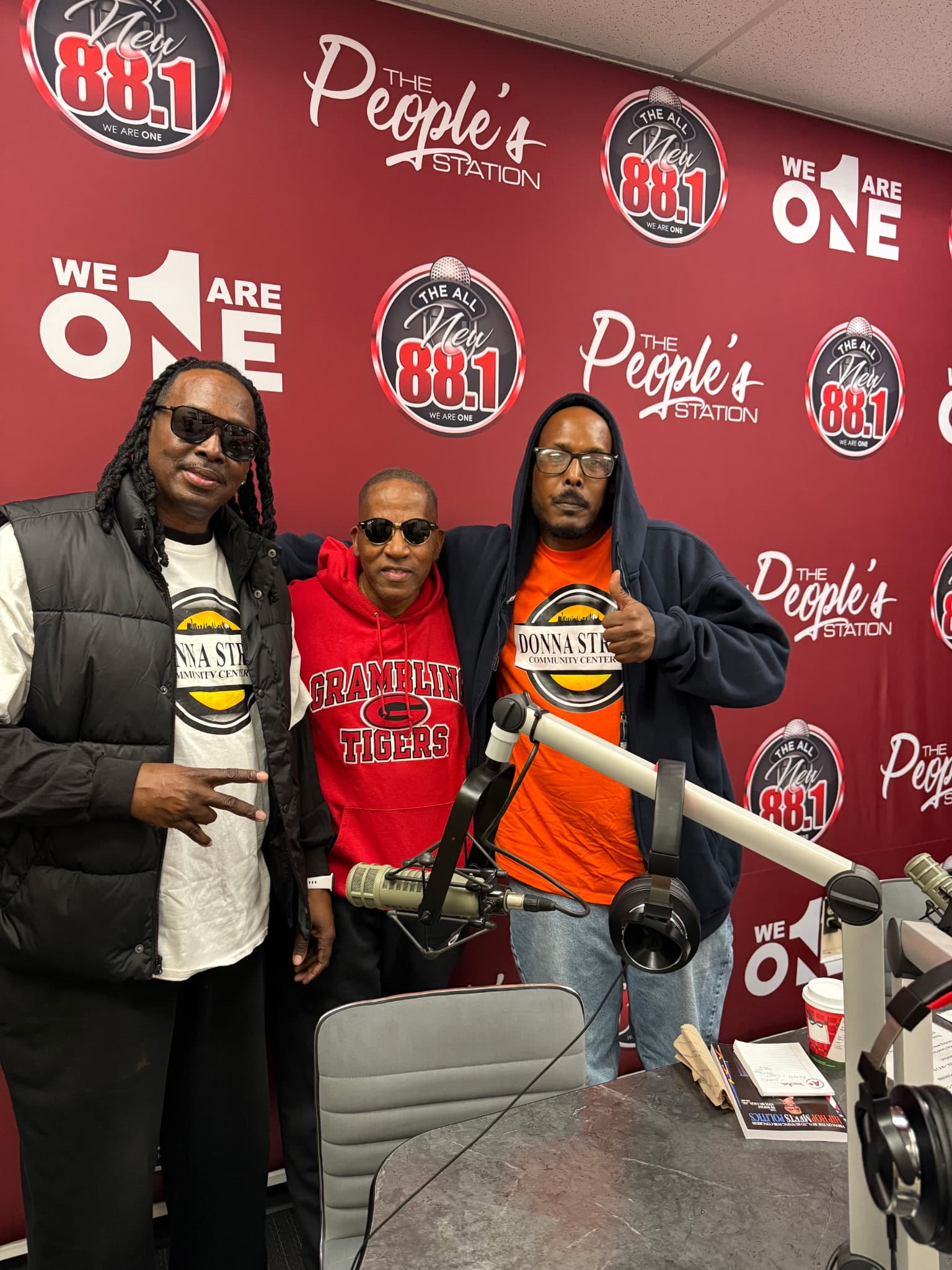 Three men pose in a radio studio against a red wall with 88.1 station logos.